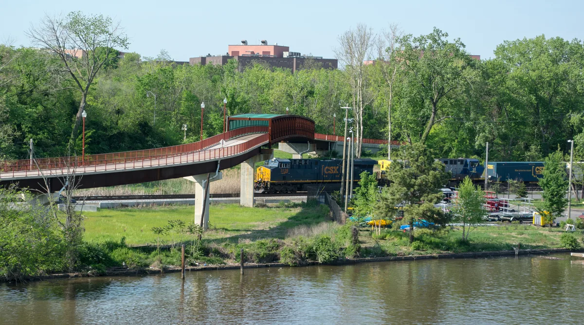 A CSX freight train passes beneath the red Anacostia Trail footbridge beside the river, framed by dense spring-green trees and brush. Calm water in the foreground reflects the shoreline while the curved bridge and tracks lead the eye through the scene.