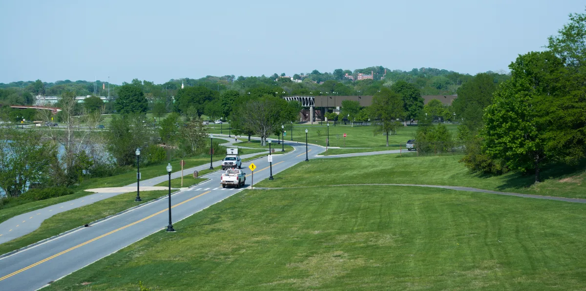 A wide view of the Anacostia Park Riverwalk shows a gently curving paved trail lined with black lamp posts and open green lawns, with the river visible at left. A few maintenance trucks sit on the road near the center, while leafy trees and a low bridge or building stretch across the hazy spring landscape in the distance.