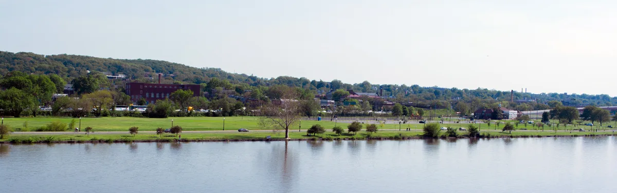 Wide view across the Anacostia River showing a calm, reflective water surface in the foreground and a strip of green parkland with scattered trees along the far bank. Low hills, a few buildings, and a bright hazy sky stretch across the background, giving the scene an open, peaceful feel.