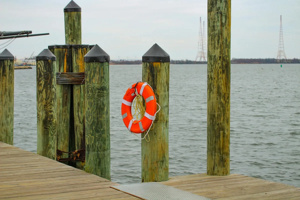 Weathered wooden dock posts line the edge of Annapolis City Dock, with an orange life ring hanging from one post and calm gray harbor water stretching behind it. In the distance, sailboat masts and a low shoreline sit under a pale overcast sky.