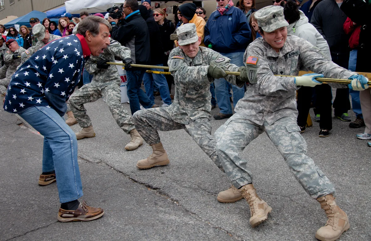 Three soldiers in camouflage and tan boots brace low in a tug-of-war, pulling hard on a thick rope against a man in a star-spangled sweater. A bundled crowd stands close behind them on the street, watching the competition in overcast daylight.
