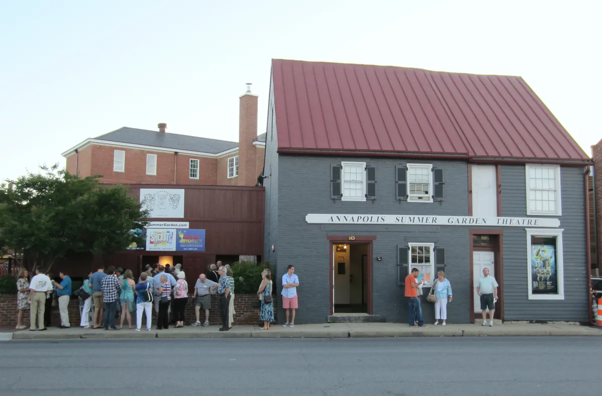 People line up outside the gray, red-roofed Annapolis Summer Garden Theatre, with a brick building and leafy tree tucked behind it. The scene feels like a warm evening street view, with small groups gathered along the sidewalk under soft light.
