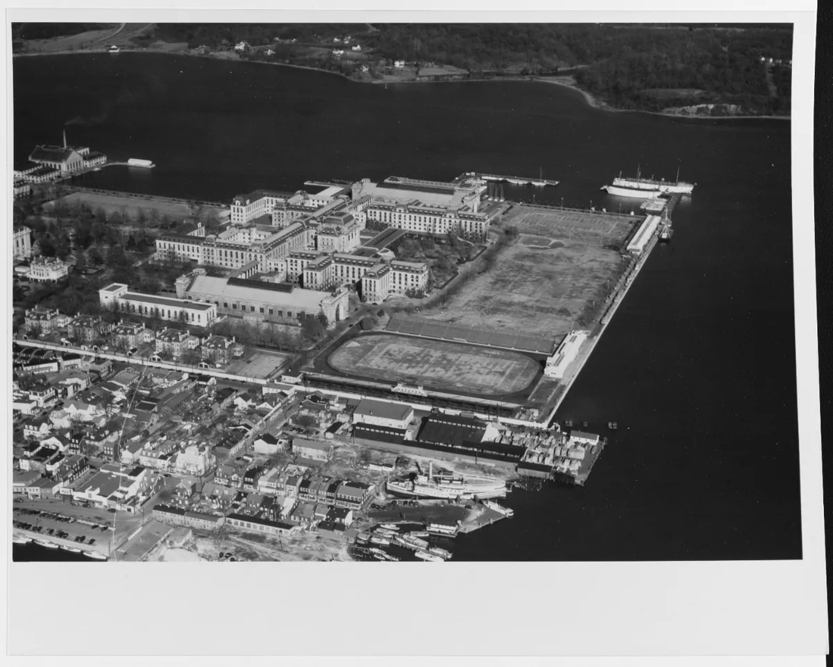 Black-and-white aerial view of Annapolis City Dock with the Maryland State House and surrounding colonial buildings clustered beside the harbor. Sailboats and a larger ship sit at the piers, while the waterfront roads, docks, and open water frame the historic downtown.