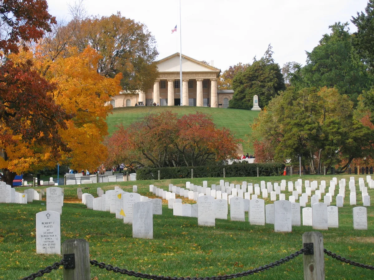 White marble headstones arc across a green hillside in Arlington National Cemetery, leading the eye up to Arlington House centered on the ridge. Autumn trees in orange, gold, and green frame the scene, with the American flag flying above the columned portico.