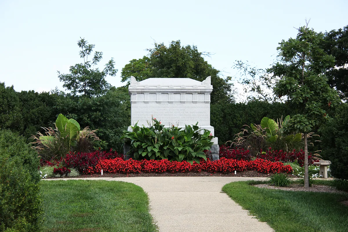 A white marble memorial stone sits centered at the end of a curving concrete path, framed by dense green shrubs and trees. Bright red and deep burgundy flower beds create a vivid border around the monument in a carefully maintained garden setting.