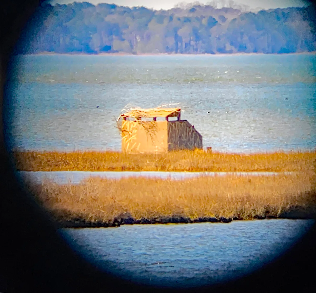 A small tan wildlife blind sits on a narrow strip of marsh grass and sand, framed by dark circular vignetting as if viewed through binoculars. Blue-gray water fills the foreground and middle distance, with a soft, wooded shoreline glowing faintly in the background light.