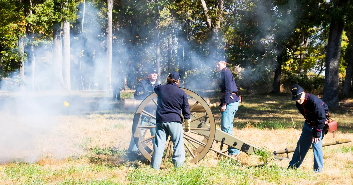 Civil War reenactors in blue uniforms work around a field cannon as white smoke drifts across a sunlit grassy clearing. Tall hardwood trees and scattered autumn leaves frame the scene, giving the battlefield a hazy morning atmosphere.
