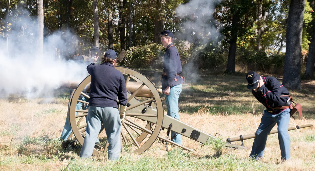 Civil War reenactors in Union uniforms fire a cannon in a grassy clearing, with thick white smoke drifting through the morning light. Tall hardwood trees frame the scene, and the cannon’s wooden wheel and long barrel stand out against the dry autumn grass.