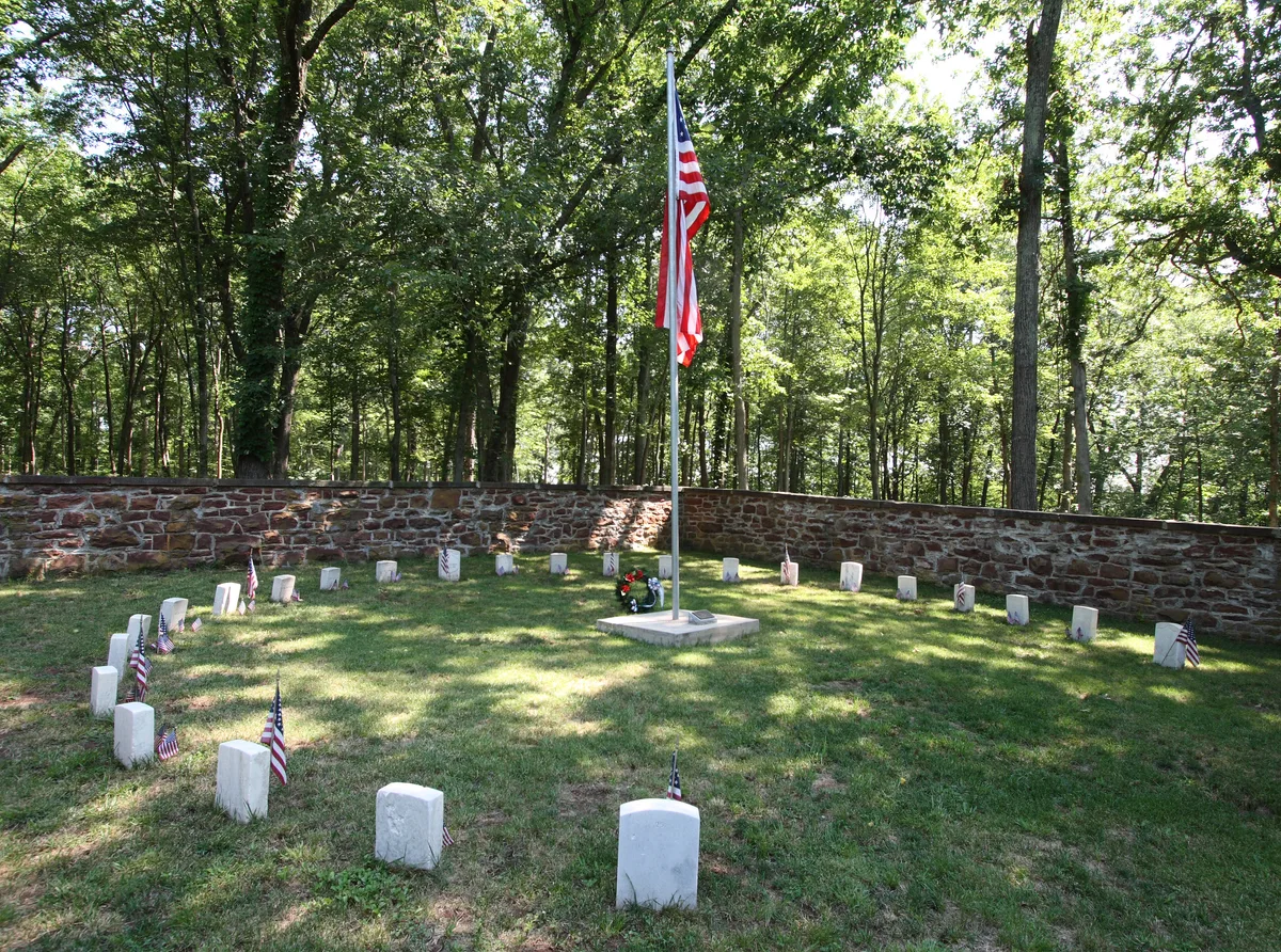 A small Civil War cemetery sits in a wooded clearing, with a ring of white grave markers surrounding a tall American flag on a concrete base. A low stone wall encircles the grassy plot, while sunlight filters through the trees onto the quiet memorial.