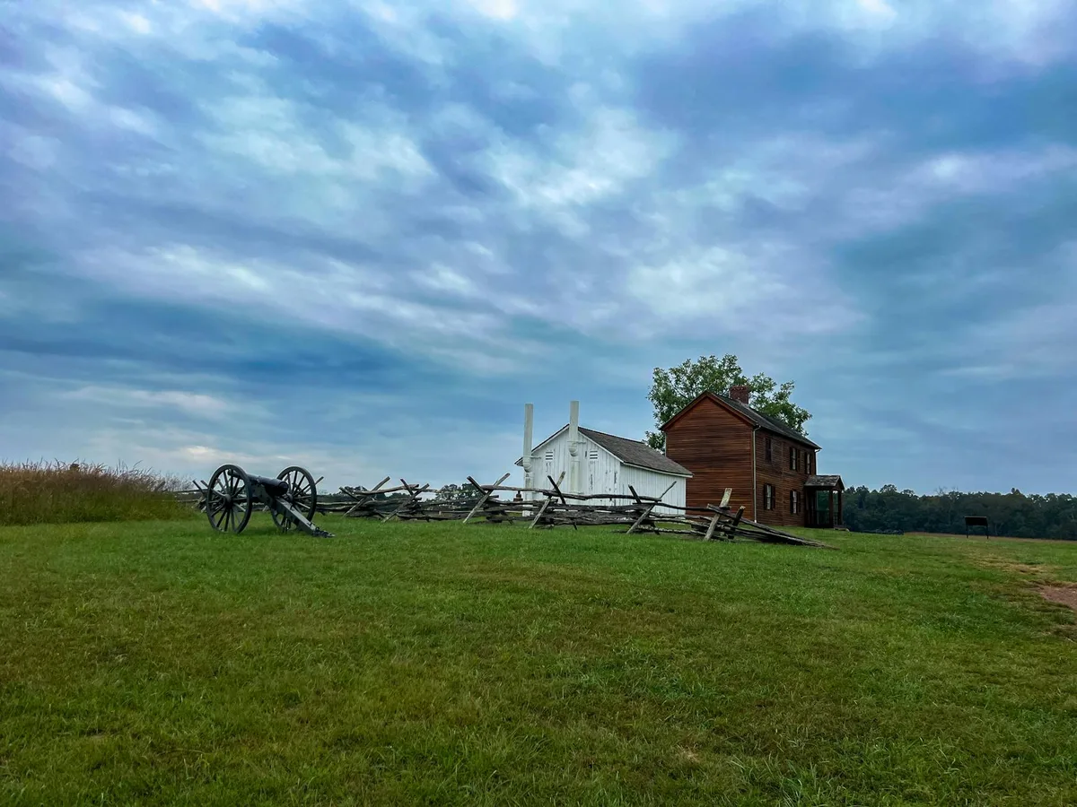 A Civil War cannon and split-rail fence sit on a grassy bluff beside two historic buildings, one white and one red-brown, under a dramatic blue-gray morning sky. The open field and distant tree line give the scene a quiet, atmospheric feel.