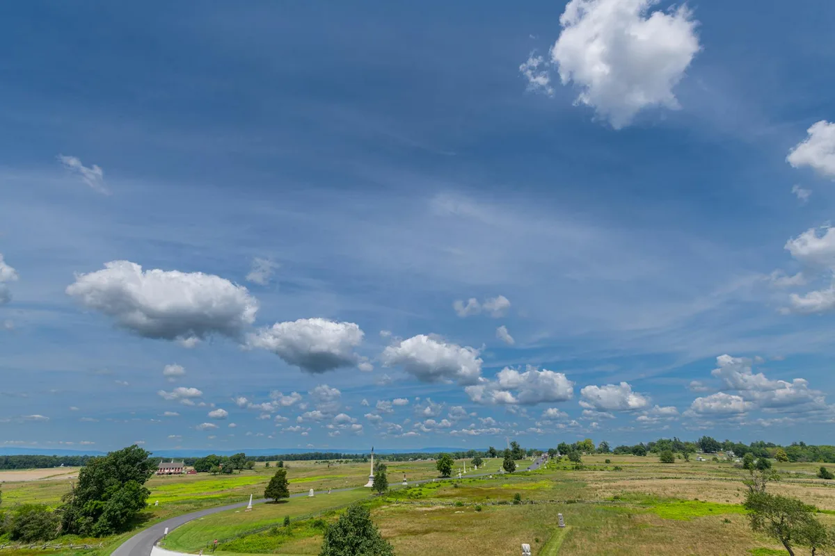 A wide view of Ball’s Bluff Battlefield shows rolling green fields and a winding paved road dotted with white memorial markers, leading toward a tall monument near the center. The bright blue sky is filled with puffy clouds, with the Potomac bluffs and tree line stretching across the horizon.
