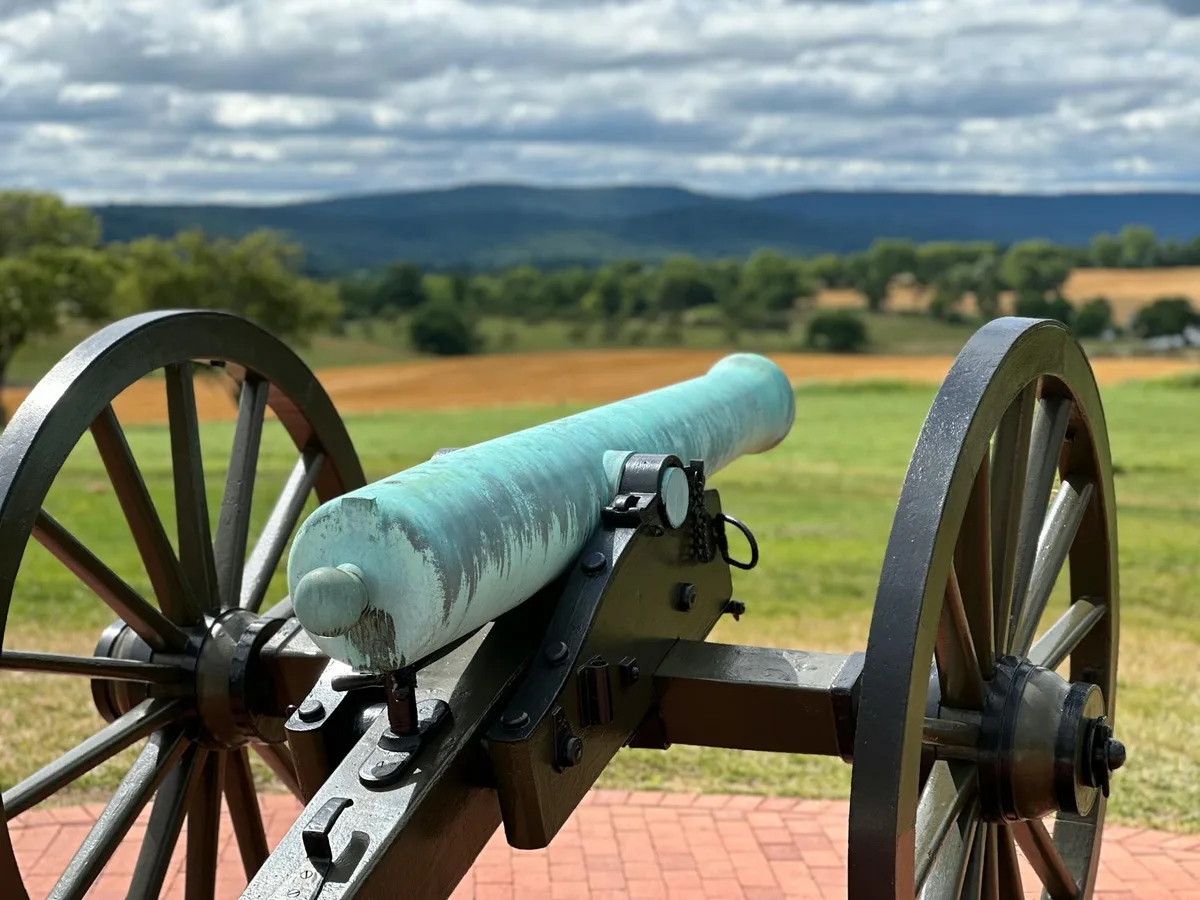 A weathered Civil War cannon sits in the foreground on a brick pad, its blue-green barrel framed by large wooden wheels. Behind it, rolling green fields and distant blue hills stretch under a heavy, cloud-filled morning sky.