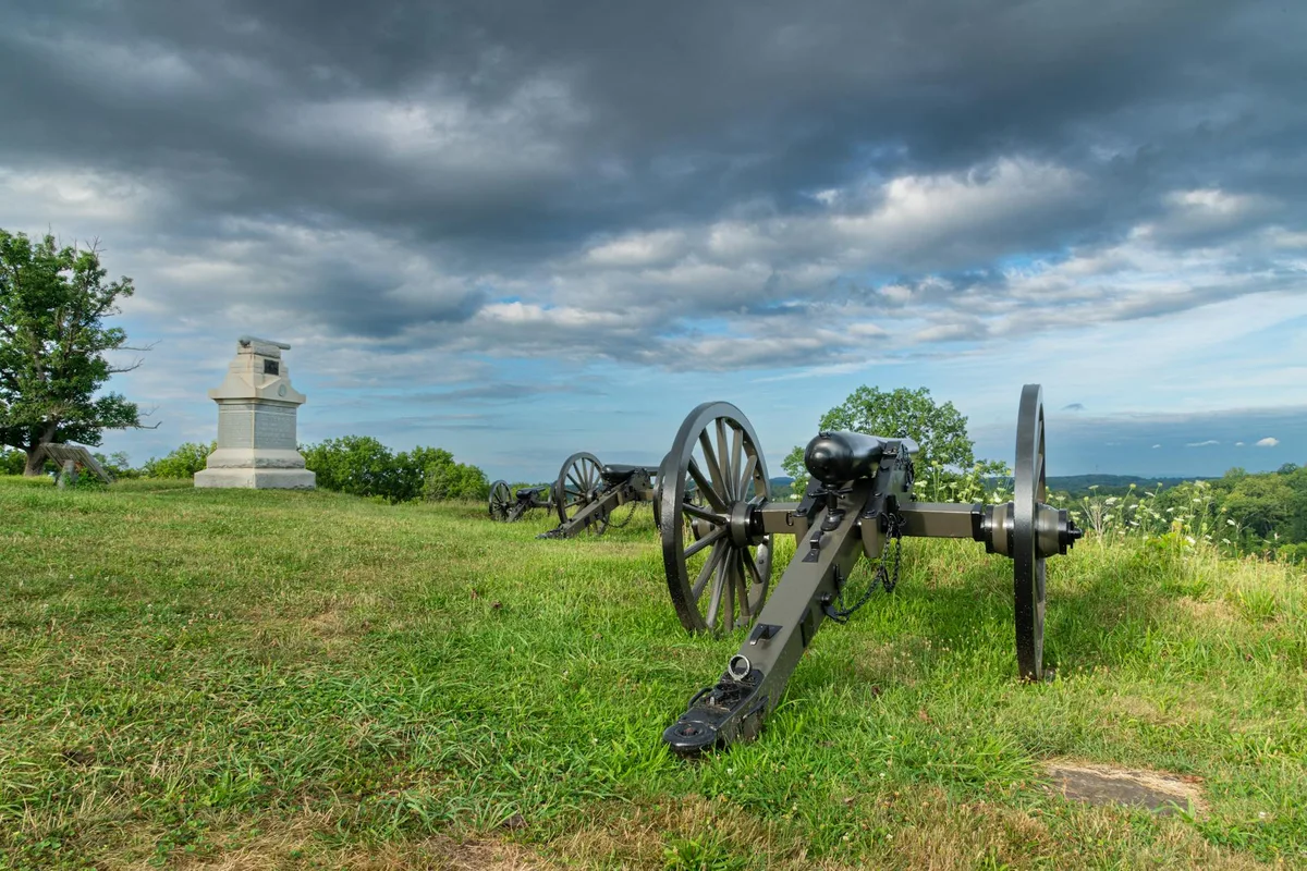 Civil War cannons line a grassy bluff beside a small stone monument, with sweeping views over the Potomac valley. Heavy gray clouds stretch across the sky, giving the scene a moody, historic feel.