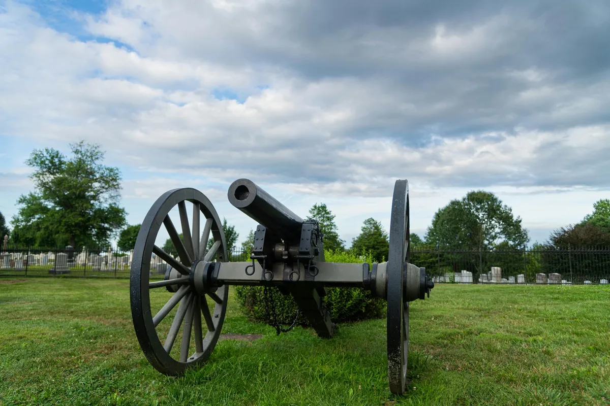 A Civil War cannon sits on a grassy bluff, framed by a low iron fence, small headstones, and scattered trees under a dramatic blanket of gray clouds. The wide, low-angle composition emphasizes the cannon’s wheels and barrel against the open sky.