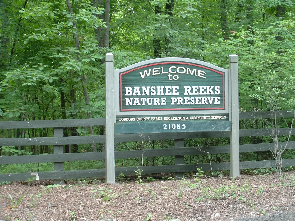 A wooded trail entrance with a large green-and-white sign reading “Welcome to Banshee Reeks Nature Preserve” stands in front of dense leafy trees and a dark split-rail fence. The foreground is a mulched roadside edge, with the preserve entrance framed by summer greenery.