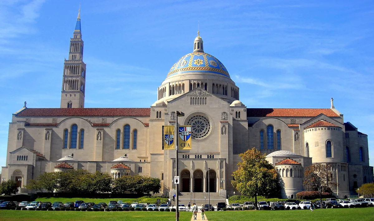 The Basilica of the National Shrine fills the frame in bright midday light, with its blue mosaic-clad dome centered above a pale stone façade and rose window. The tall bell tower rises on the left, while red-tiled roofs, arched windows, and a green lawn with parked cars stretch across the foreground under a clear blue sky.