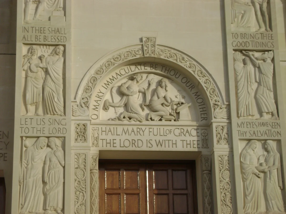 A carved stone entrance inside the basilica is framed by an arched lintel and shallow relief figures, with the words “Hail Mary full of grace, the Lord is with thee” across the top. Warm light falls on the cream-colored walls and wooden doors below, highlighting the ornate Byzantine-style details and scripture panels on either side.