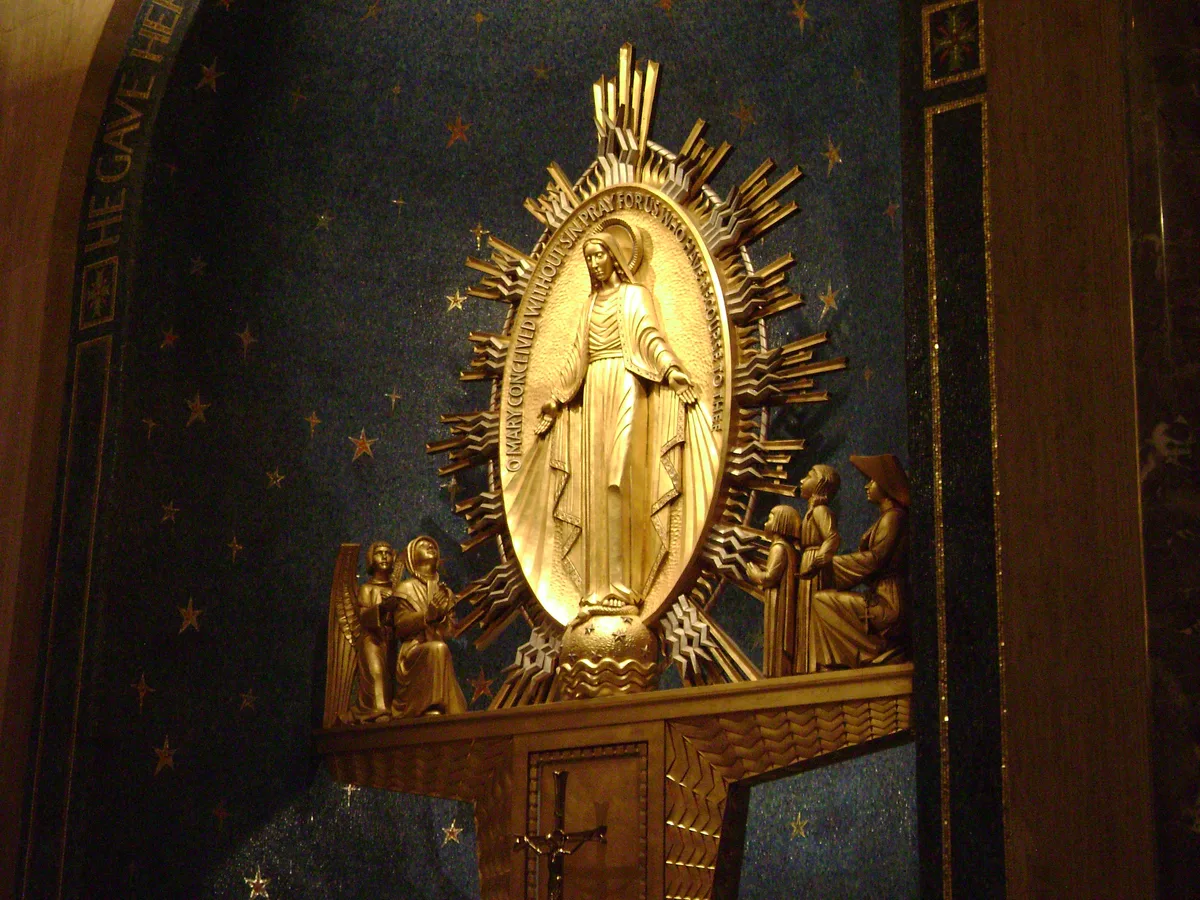 A gilded relief of the Virgin Mary stands in an oval halo against a deep blue mosaic wall, surrounded by radiating bronze rays and small star motifs. Smaller figures kneel at the base near a wooden altar panel, with dark chapel wood framing the glowing devotional scene.