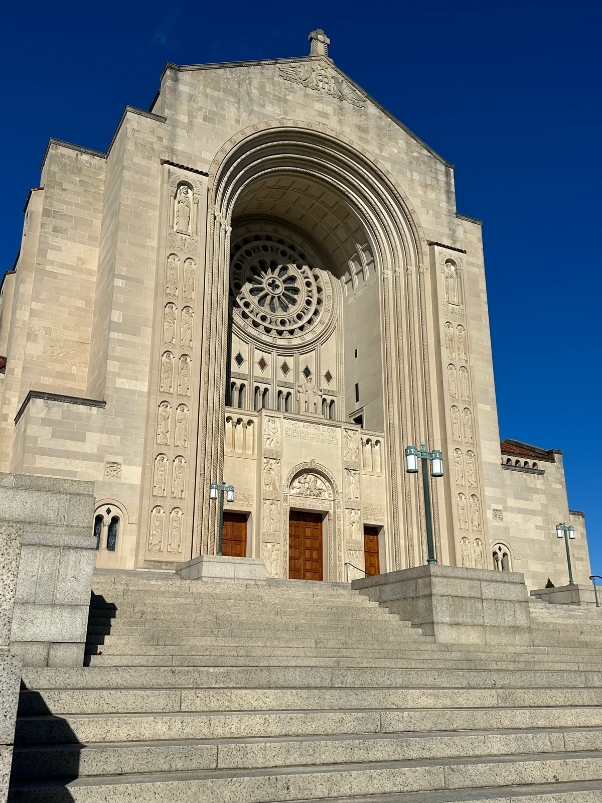 Grand stone steps lead up to the basilica’s monumental main entrance, framed by a tall arched portal, carved saints, and a large circular rose window. The pale limestone façade glows in bright midday light against a deep blue sky.