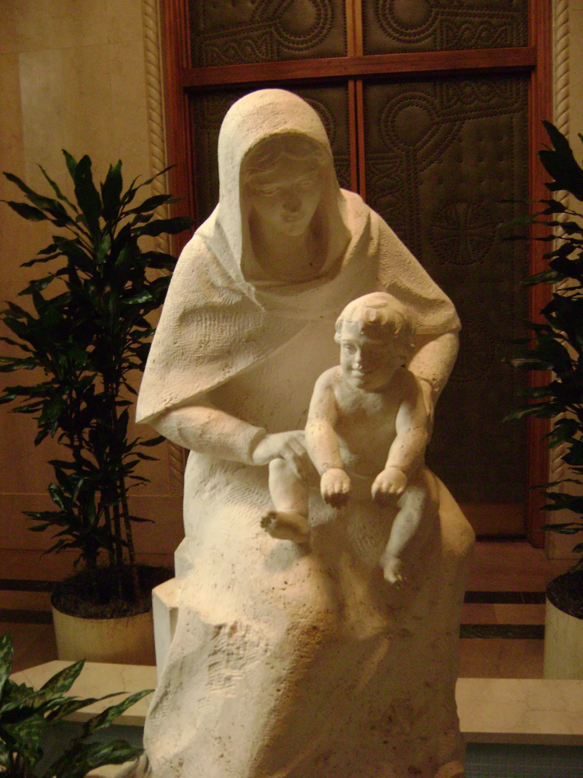 A softly lit white stone statue of a veiled woman holding a small child stands in a shrine alcove, framed by potted plants and a wood-trimmed window behind her. The warm interior light highlights the rough texture of the sculpture and the serene, devotional pose.