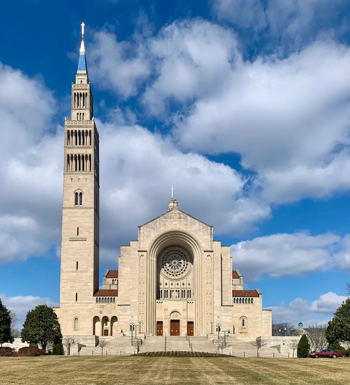 The Basilica of the National Shrine rises in pale stone beneath a bright blue sky, with its tall bell tower capped by a gold cross and blue spire beside a grand arched entrance and circular rose window. Wide steps, low red-tiled rooflines, and a broad winter-brown lawn frame the symmetrical façade.