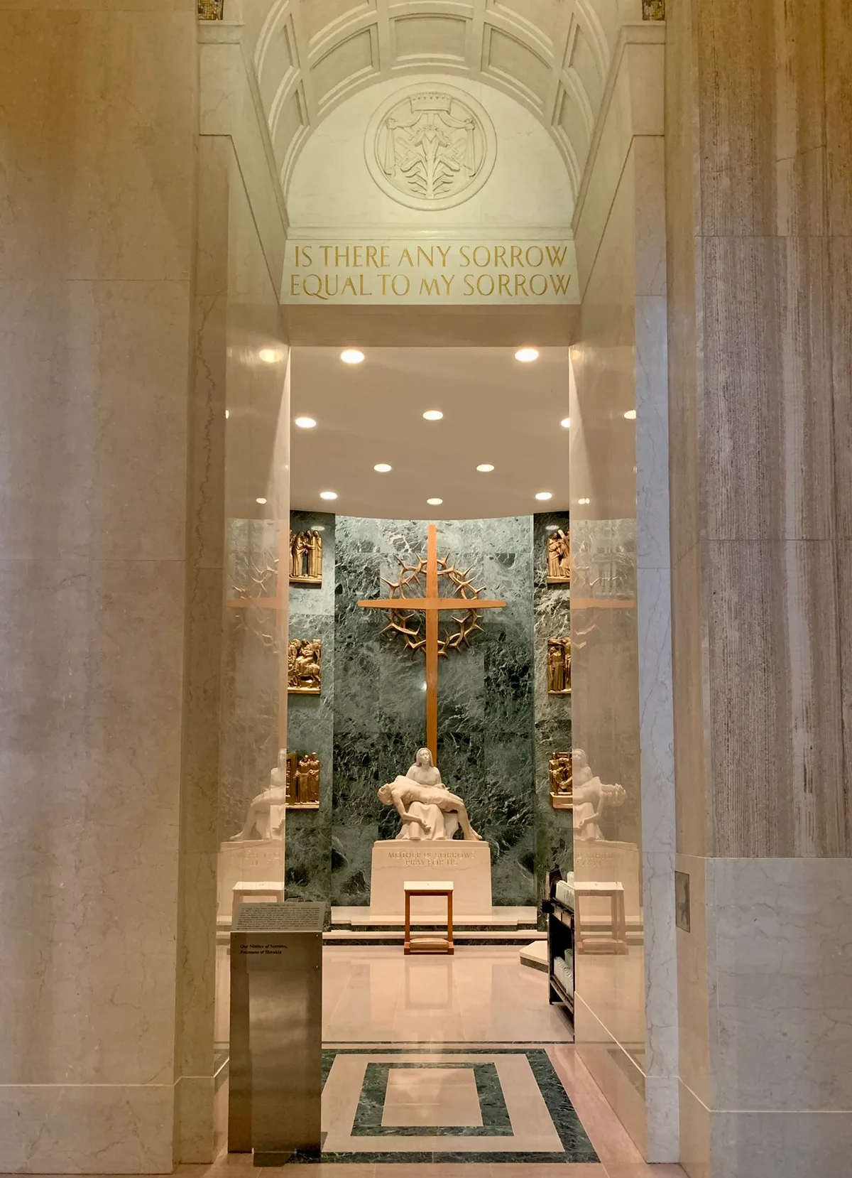 A marble chapel niche glows under recessed lights, with a gold cross centered against a dark green stone backdrop and a sculpted Pietà beneath it. Above the doorway, gold lettering reads “IS THERE ANY SORROW EQUAL TO MY SORROW,” framed by pale stone arches and subtle religious reliefs.
