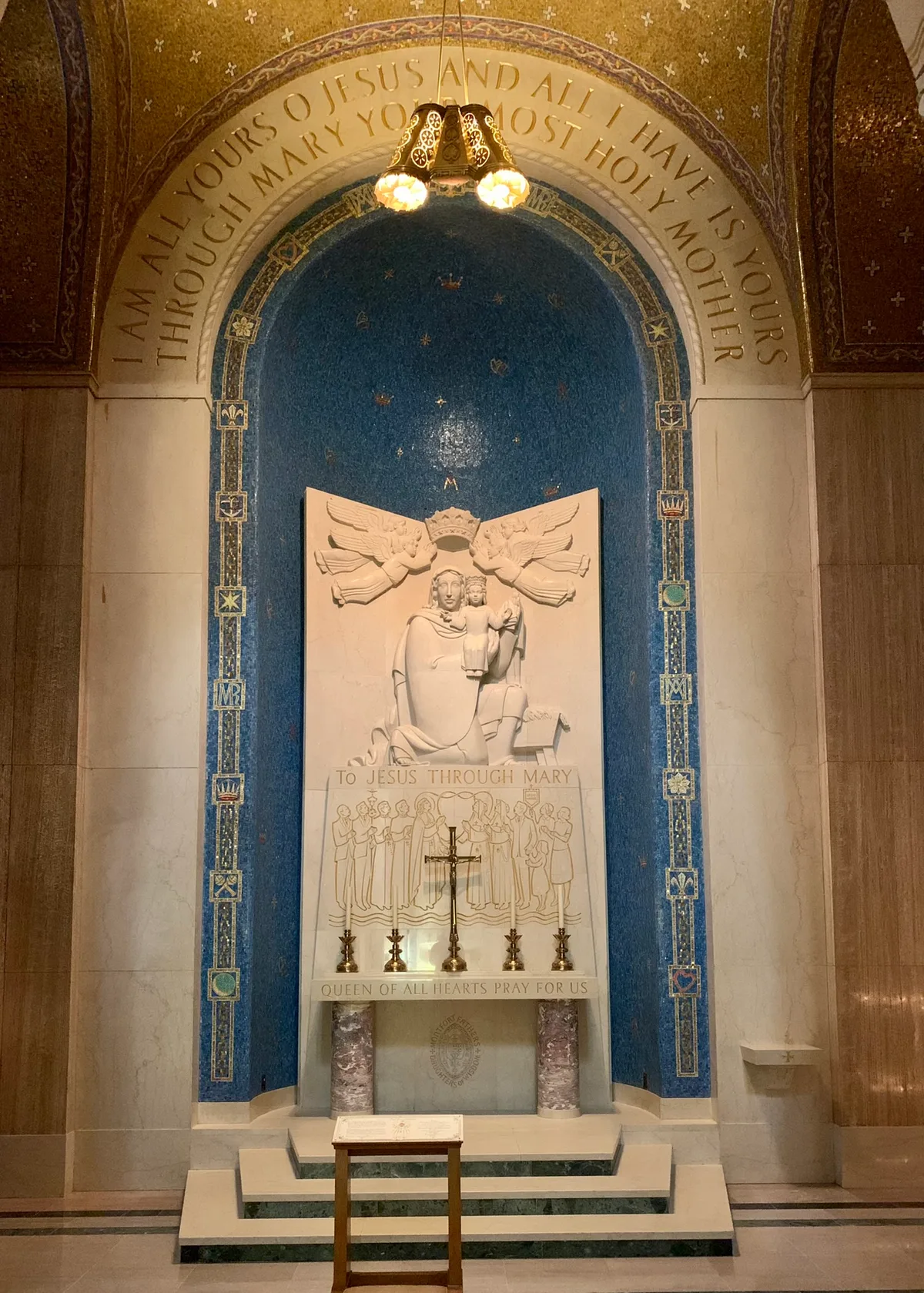 A marble prayer alcove glows under a small hanging lamp, framed by a deep blue mosaic arch and gold lettering reading “I AM ALL YOURS… THROUGH MARY MOST HOLY MOTHER.” At the center, a white relief of the Madonna and Child sits above a line of candles and the inscription “TO JESUS THROUGH MARY,” with the altar steps and stand in the foreground.