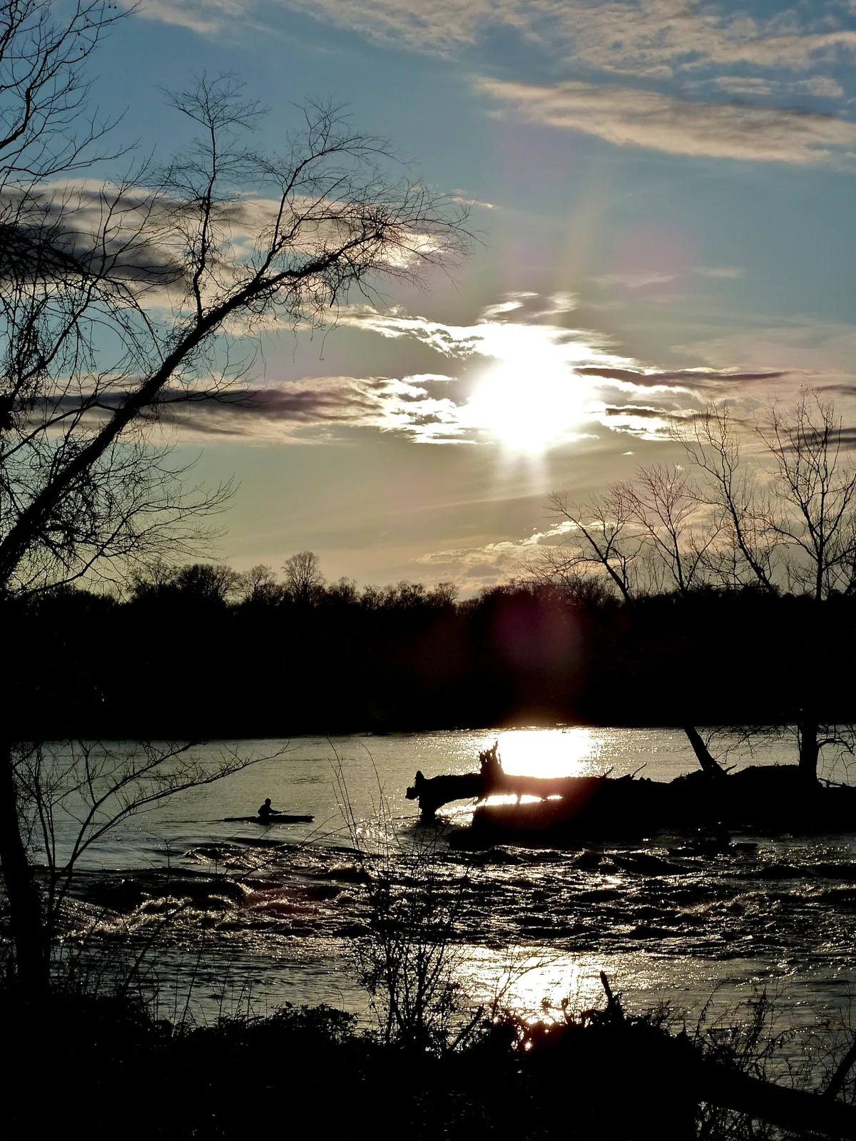 Golden-hour sun breaks through thin clouds over the James River, silhouetting leafless trees and a lone kayaker against shimmering rapids. The water glows around dark rocks and riverbank debris, creating a dramatic backlit scene.