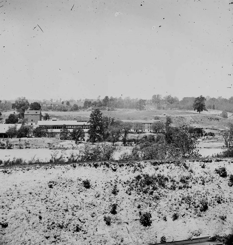 A wide view across Belle Isle shows the river in the foreground and a low industrial complex with trees and open ground on the island, under a bright, hazy sky. The scene feels sparse and rugged, with the hillside and shoreline stretching back into the distance.