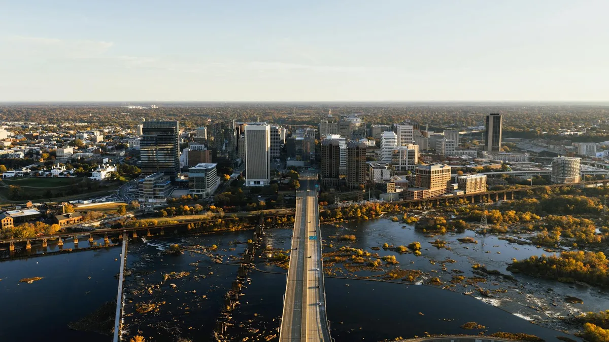 A long pedestrian bridge leads from Belle Isle toward downtown Richmond, centered over the James River’s dark rapids and scattered rocky shallows. Golden-hour light warms the skyline and autumn trees lining the riverbanks.