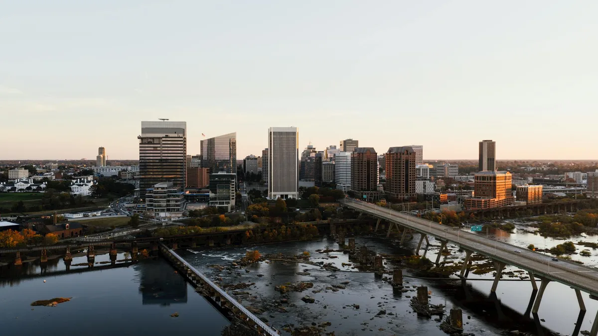Downtown Richmond’s skyline glows in soft golden light above the James River, with the pedestrian bridge stretching diagonally across the frame. Dark rippling water, scattered river rocks, and the remains of old concrete piers and ruins line the foreground.