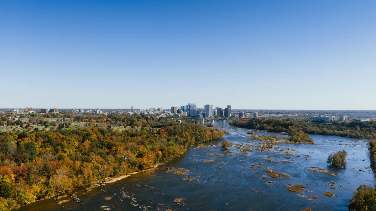 Wide view over the James River from Belle Isle, with autumn-colored trees lining the island and rocky rapids flowing past toward the downtown Richmond skyline. The clear blue sky and distant bridge create a bright, open landscape framed by water and city buildings.