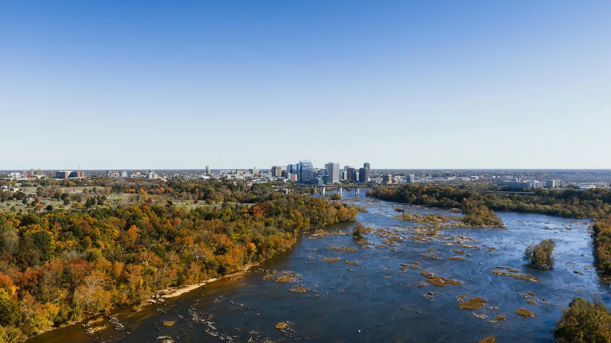 A wide aerial view shows the James River flowing past Belle Isle, with rocky rapids and low islands in the foreground and Richmond’s downtown skyline rising in the distance. Autumn trees in greens, golds, and rust line the riverbanks under a clear blue sky.