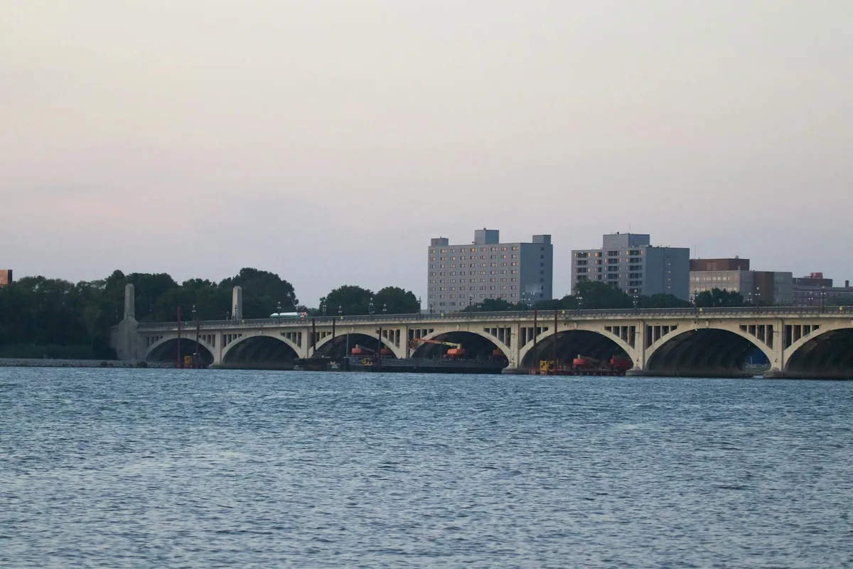 A wide view across the James River shows the cream-colored stone arches of a bridge with downtown Richmond buildings rising behind it. Calm blue water fills the foreground under a pale evening sky, with the scene softly lit at golden hour.