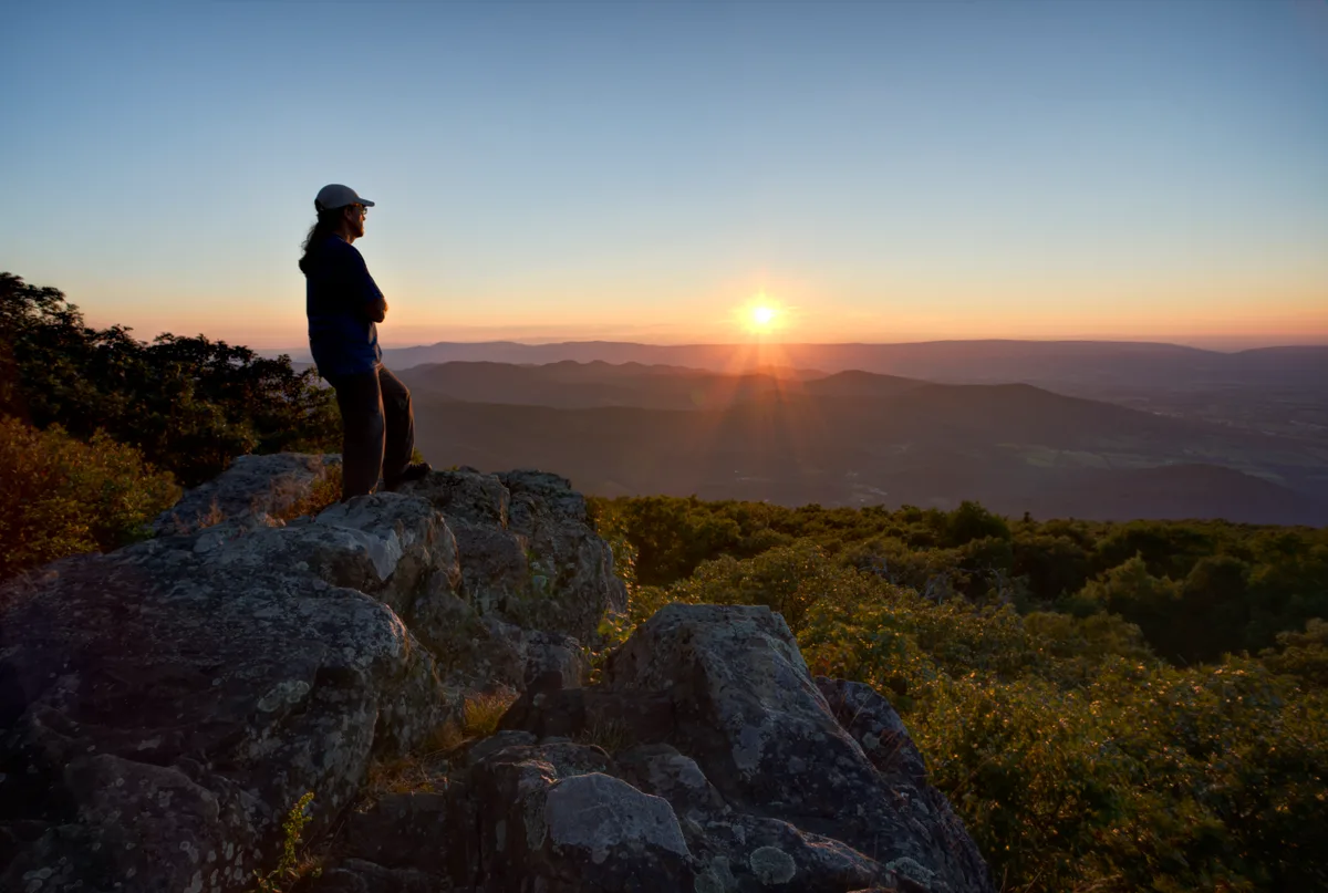 A hiker stands silhouetted on jagged quartzite boulders at Blackrock Summit, watching the sun rise over layered blue ridges in Shenandoah National Park. Warm orange light spills across the horizon while the rocky foreground and surrounding treetops stay in deep shadow.