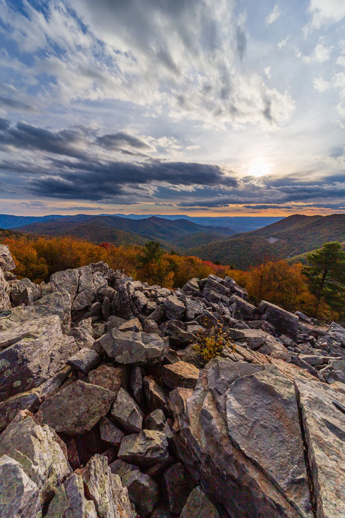 Jagged quartzite boulders fill the foreground of Blackrock Summit, opening to layered Shenandoah ridgelines and a valley glowing with autumn color. Above, dramatic streaked clouds frame the low sun and a thin band of warm light on the horizon.