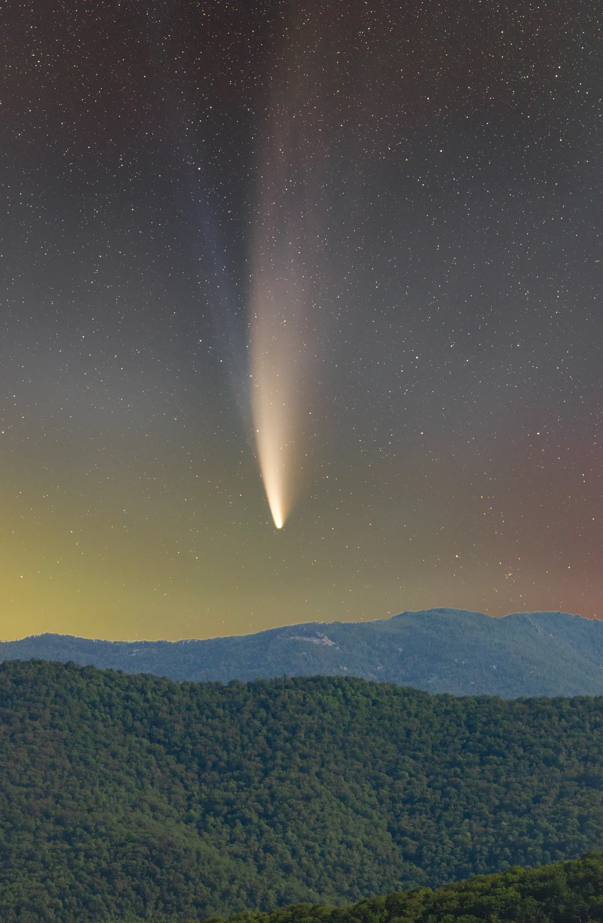 A bright comet with a glowing tail rises into a star-filled night sky above layered blue-green Shenandoah ridges. The scene has a faint yellow-green glow near the horizon, with dark forested mountains filling the lower half of the frame.