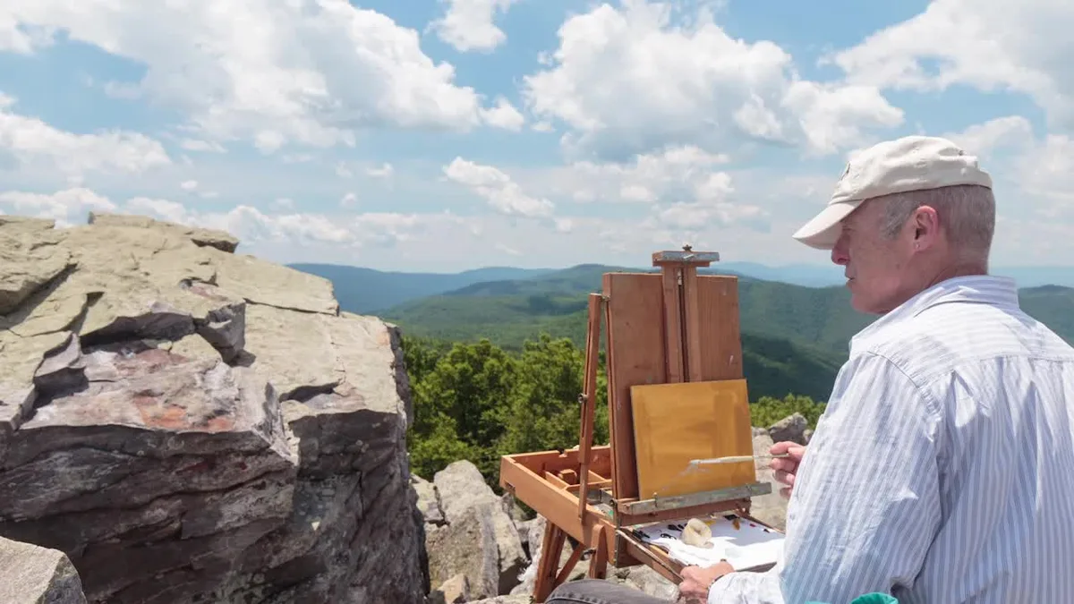 A man in a white cap paints at an easel perched on Blackrock Summit, with pale quartzite boulders in the foreground and layered blue-green Shenandoah ridges stretching under a bright, cloud-filled sky.