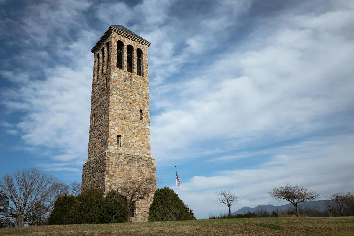 A tall stone observation tower rises from the bare quartzite summit, its rough masonry and dark roof set against a wide sky of soft blue and drifting clouds. Low shrubs and a few leafless trees dot the foreground, with distant mountain ridges barely visible along the horizon.
