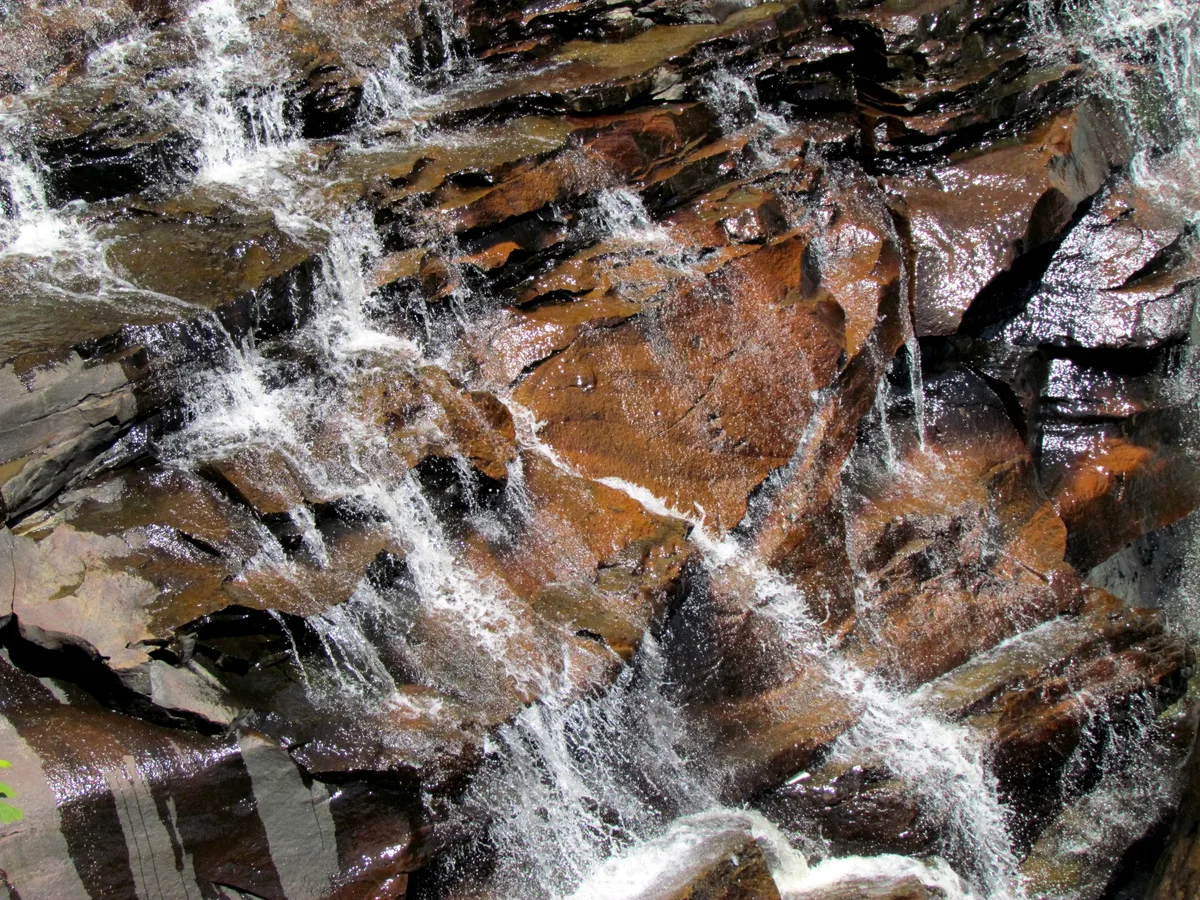 Close-up of Blackwater Falls water spilling in thin white ribbons over dark, wet sandstone, with amber-brown rock glowing in the sunlight. The frame focuses on the textured cascade and glossy ledges rather than the full falls.
