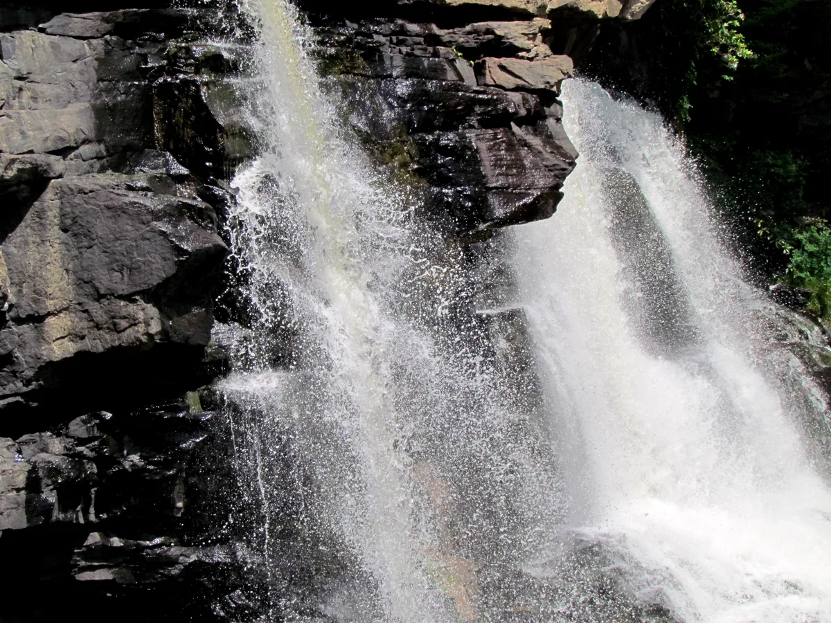 Close-up of Blackwater Falls spilling in two bright white curtains over dark sandstone ledges, with mist and spray catching the morning light. The frame is filled with the textured rock face and churning water, creating a dramatic, high-contrast view of the waterfall.