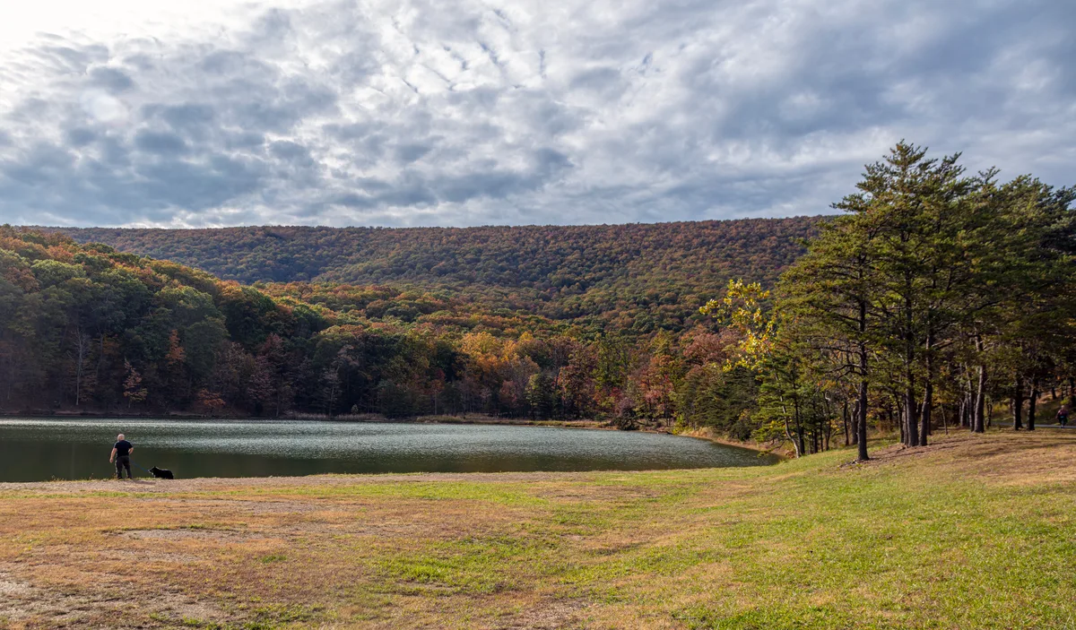 A calm upper lake curves through a grassy shoreline, with one person and a dog standing at the water’s edge beneath a wide, cloud-filled morning sky. Forested hills in early fall color rise behind the lake, and tall pines frame the right side of the view.