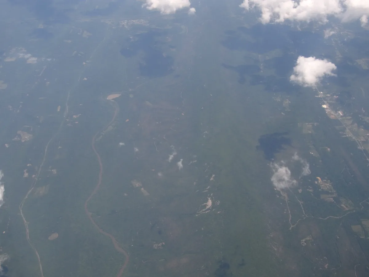 A hazy aerial view shows a broad, forested mountain ridge with a winding river cutting through the valleys below. Scattered white clouds cast soft shadows over the dark green landscape, giving the scene a misty morning feel.