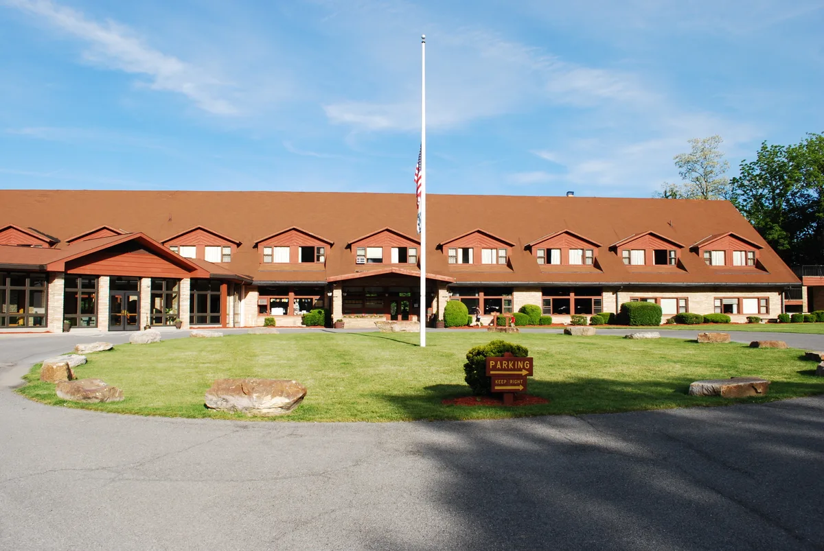 The Cacapon Resort State Park lodge stretches across the frame with a long brown roof, dormer windows, and a stone-and-wood facade centered on a tall flagpole. A neatly trimmed lawn with scattered boulders and a small parking sign sits in front under a clear morning sky.