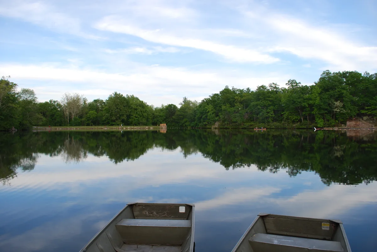 Two aluminum rowboats sit at the edge of a glassy lake, with dense green trees and a low stone shoreline reflected in the still water. Soft morning light and streaky clouds create a calm, mirror-like Appalachian scene.