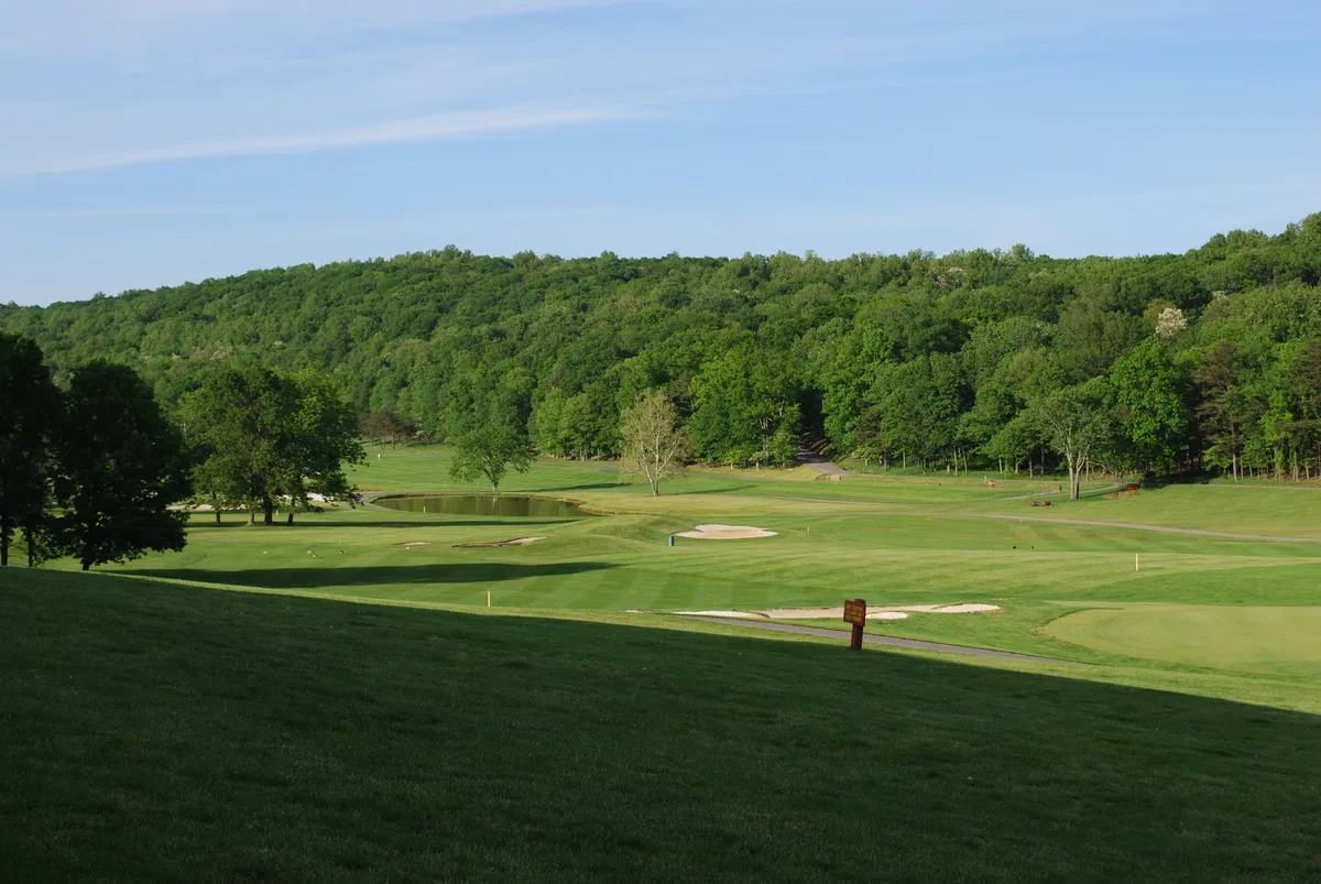 Rolling green golf fairways and sand bunkers stretch across a gently sloping valley, with a small reflective pond near the center and dense forested ridges rising in the background. Bright morning light and a clear blue sky give the scene a calm Appalachian resort feel.