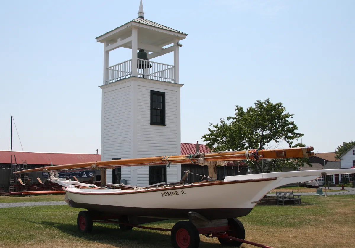 A white wooden lighthouse tower rises behind a restored log canoe on a wheeled cradle, with its long mast stretching across the frame. The scene sits on a grassy museum lawn under a clear blue sky, with a few historic waterfront buildings in the background.
