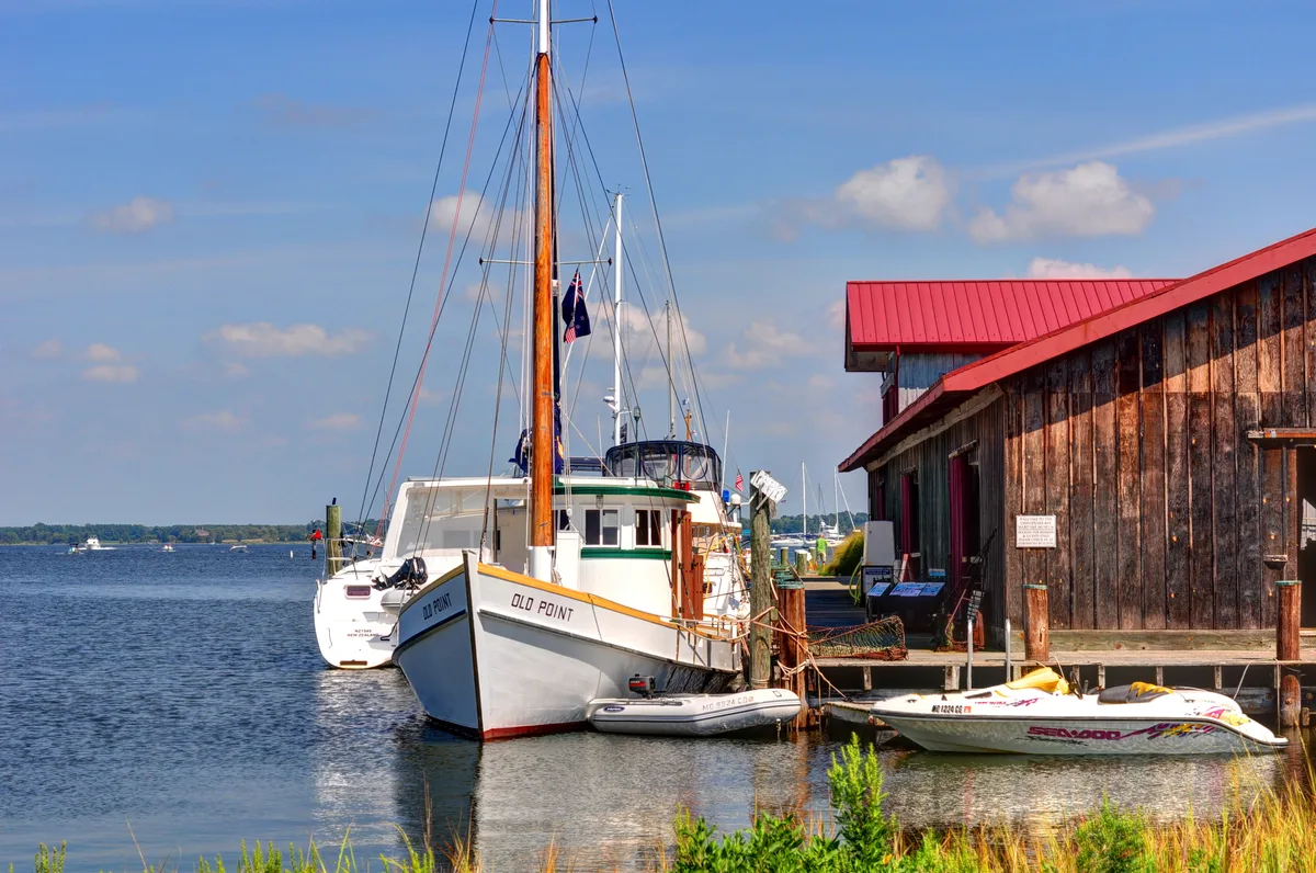 Historic workboats and sailboats are tied up along the Chesapeake Bay Maritime Museum dock, with the white bow of the “Old Point” centered beside a weathered wooden building topped by a bright red roof. Calm blue water and a wide open bay fill the left side of the frame under a clear sky.