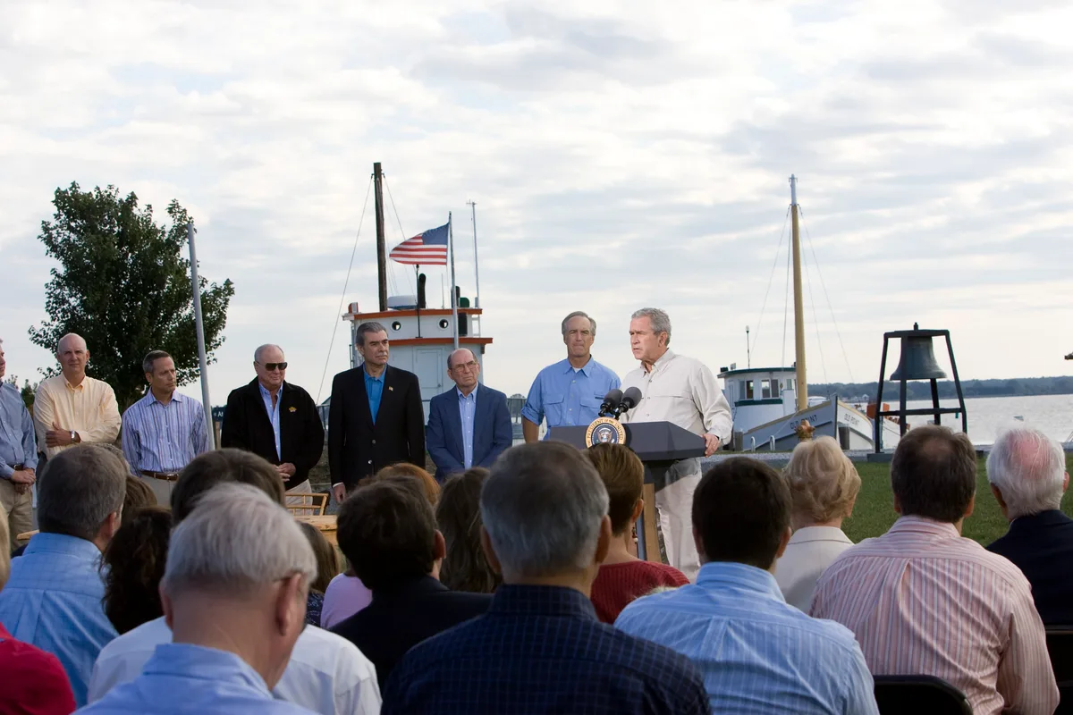 A waterfront ceremony unfolds at the Chesapeake Bay Maritime Museum, with a speaker at a podium facing a seated crowd and officials lined up beside historic boats. In the background, a workboat and a small bell structure sit on the grassy harbor edge under a bright, cloud-filled sky.