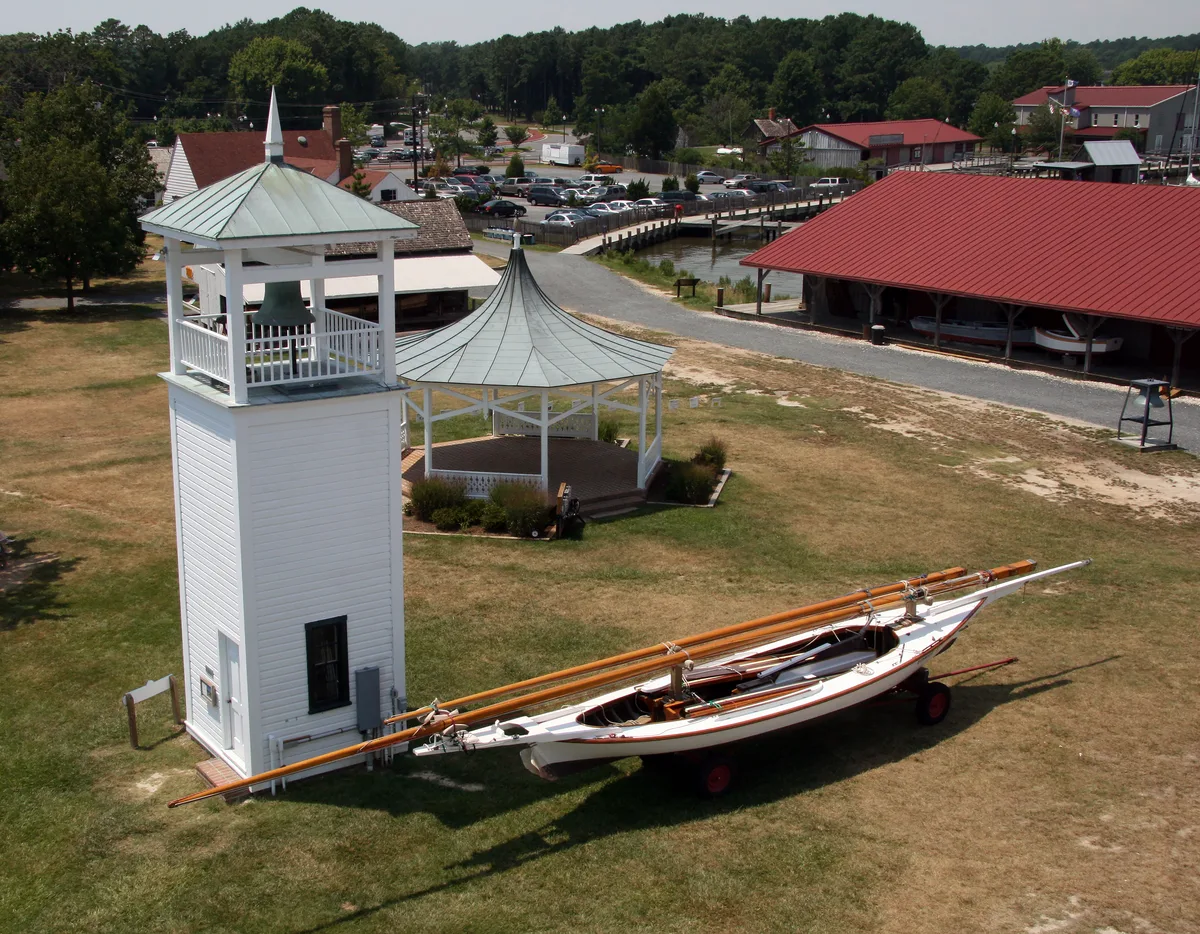 A white maritime bell tower and adjacent gazebo stand on a grassy museum lawn, with a long wooden workboat on a trailer in the foreground. Red-roofed boathouses, a dock, and parked cars line the water beyond, with trees filling the background.